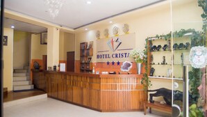 Een hotel receptiebalie met een houten toonbank, een kroonluchter en een bord dat aan de muur hangt met de tekst 'HOTEL CRISTAL'.