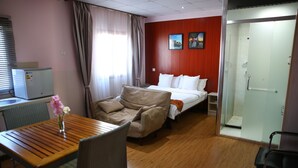 Hotelkamer met een houten tafel, een bank, een bed met witte lakens, een glazen douchedeur en een rode wand met twee ingelijste foto's.