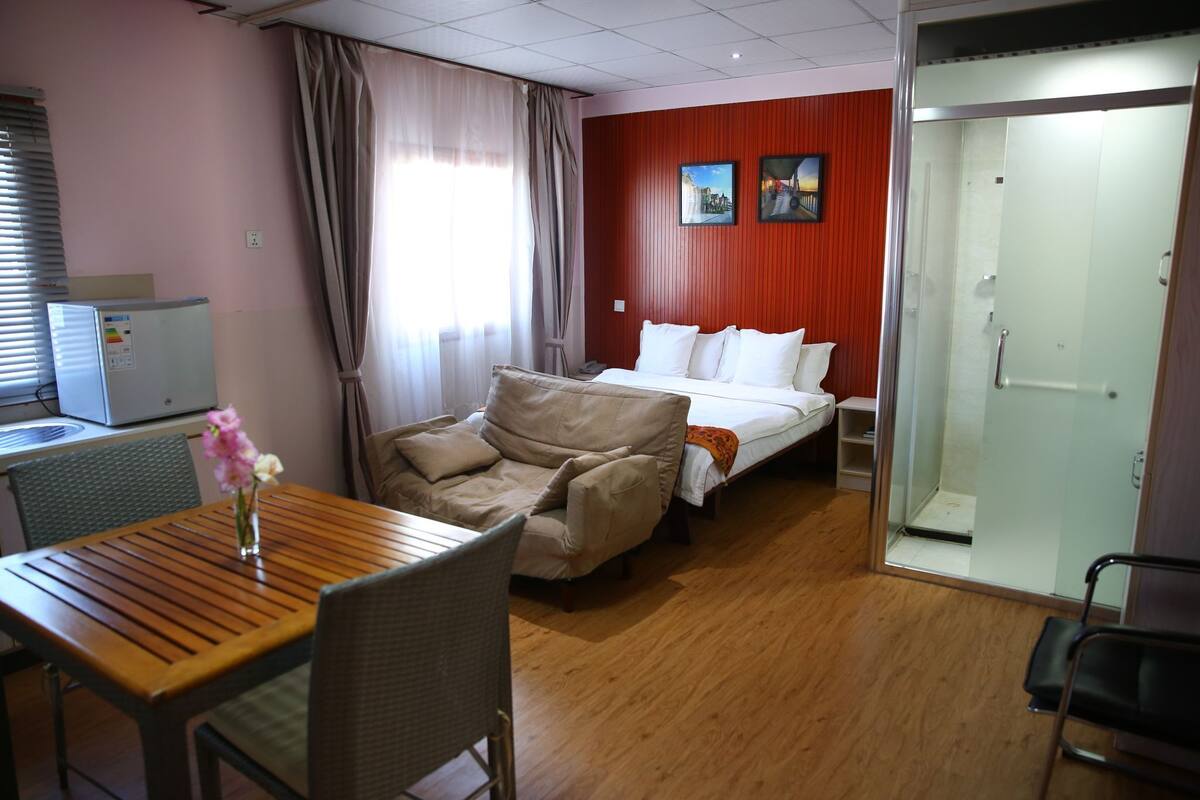 A hotel room with a wooden table, a sofa, a bed with white bedding, a glass shower door, and a red wall with two framed pictures.