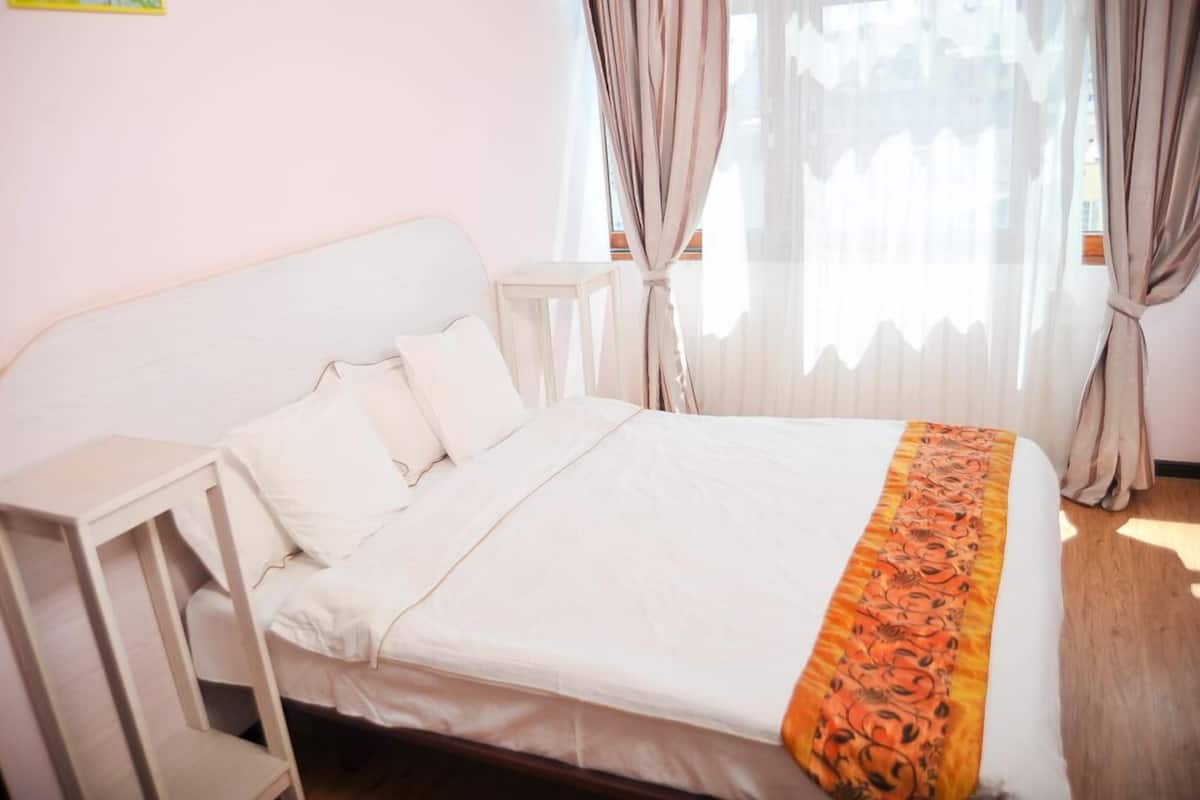 A neatly made bed with white linens and a decorative orange and black blanket. There is a small bedside table and a window with curtains.