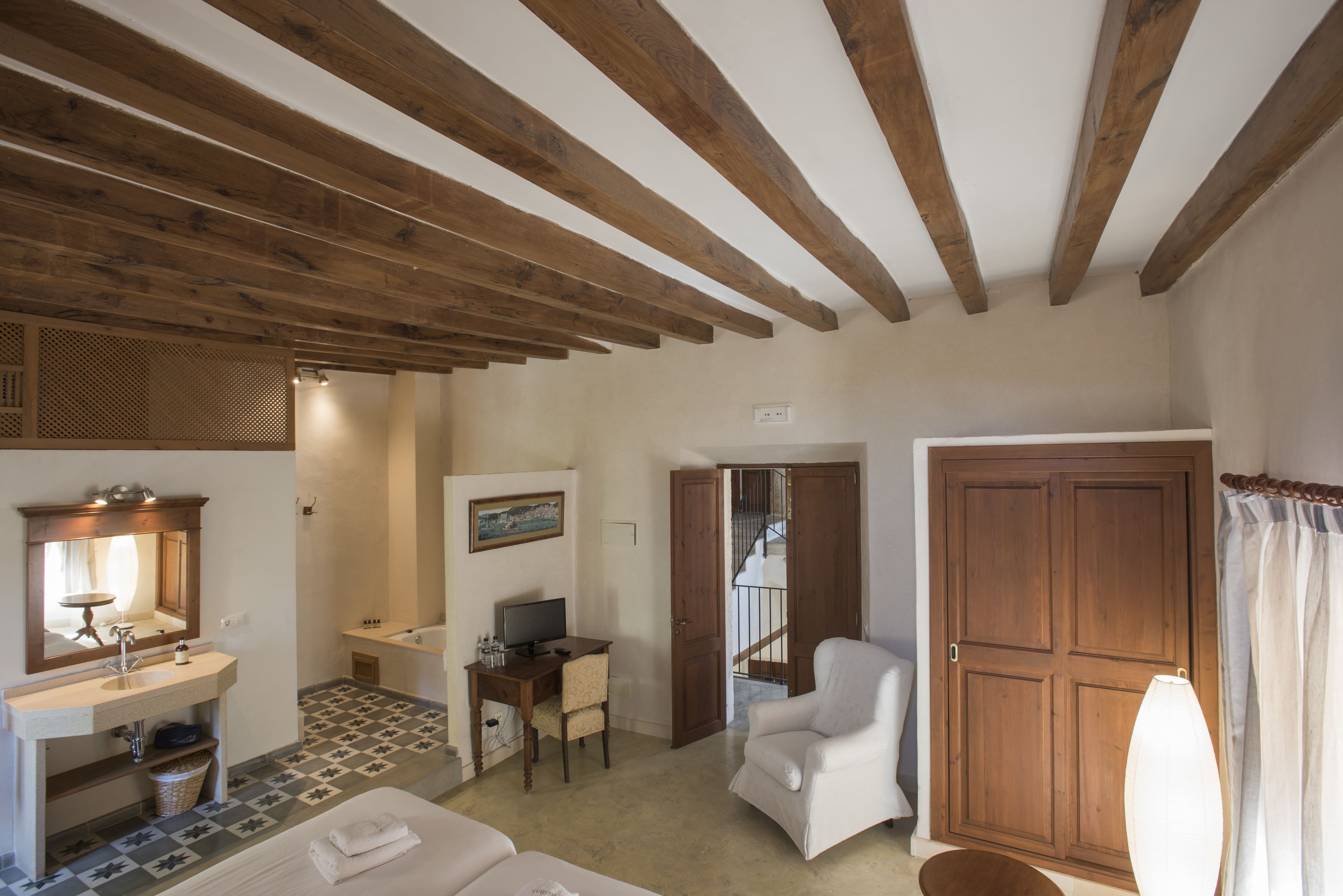 A room with a wooden ceiling, a bed, a desk with a chair, a white armchair, and a large wooden door.