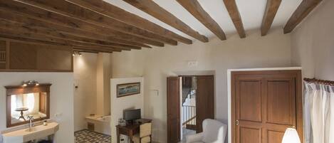 A room with a wooden ceiling, a bed, a desk with a chair, a white armchair, and a large wooden door.
