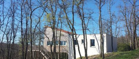 Una casa moderna con fachada blanca y tejado rojo, rodeada de árboles altos.