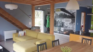 A cozy living room with a yellow sofa, wooden beams, a dining table, and a kitchen area with a cityscape mural. - Modern wooden house with tree terrace and private heated pool (Luzech)