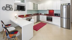 Une cuisine moderne avec des armoires blanches, un réfrigérateur en acier inoxydable, un four encastré et un four à micro-ondes. Un tapis rouge recouvre le sol, et un petit îlot central est équipé d’un évier et d’une chaise.