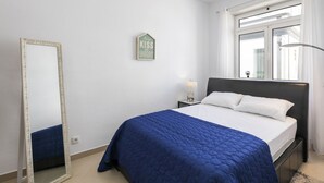 Une chambre à coucher avec un grand lit, un miroir, une table de chevet avec une lampe, une fenêtre avec des rideaux et un panneau fixé au mur.