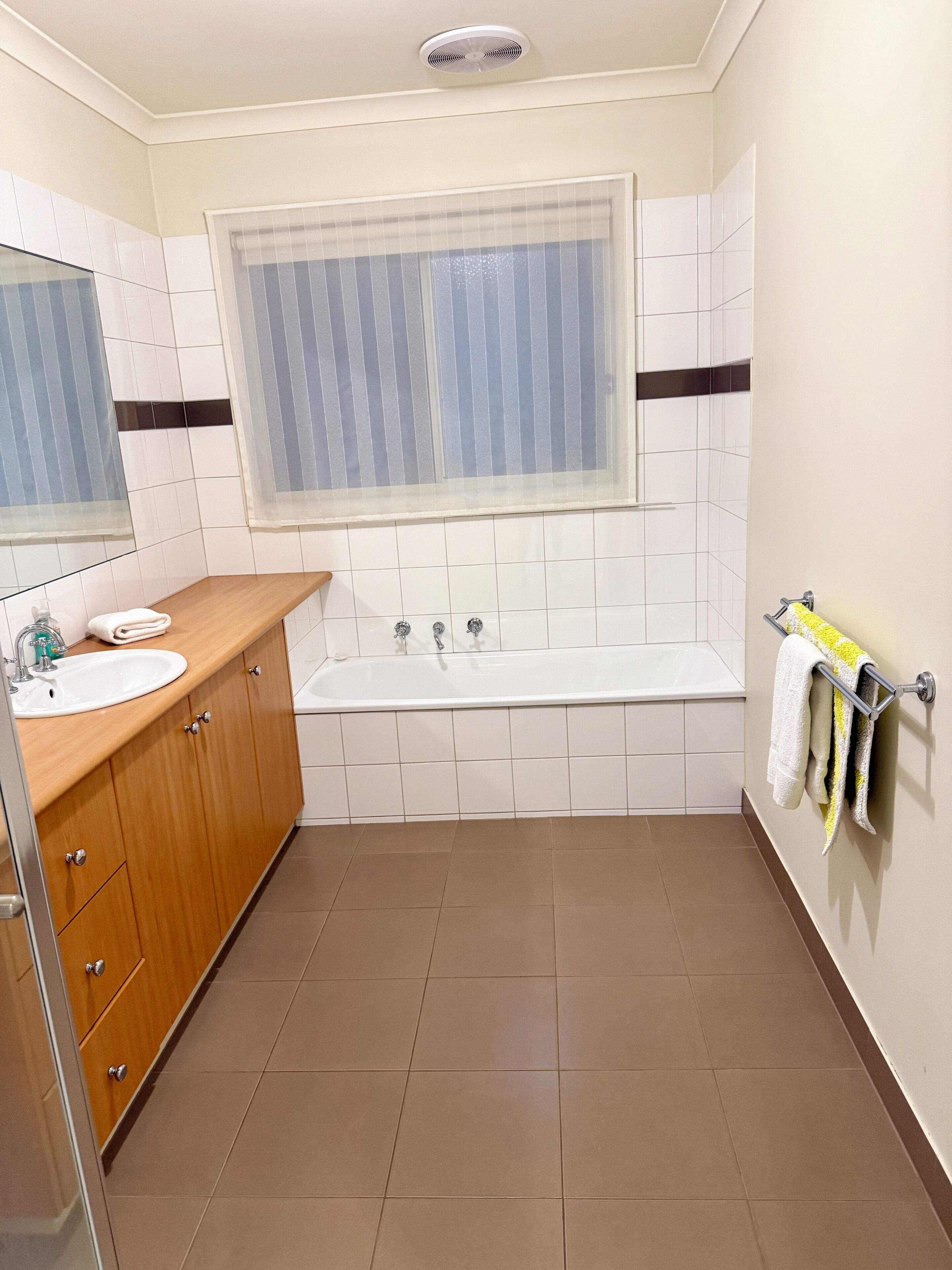 A modern bathroom with a wooden vanity, a white sink, a bathtub, and a window with blinds.