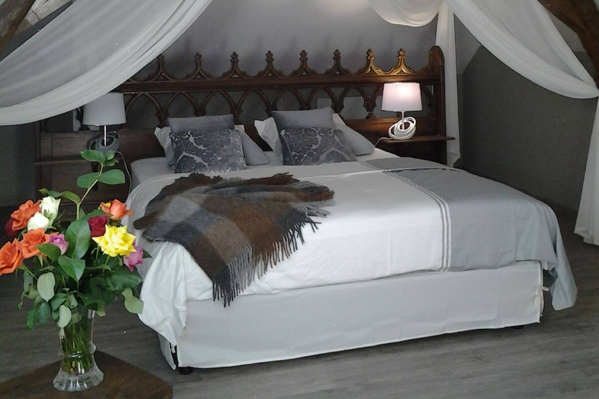 A bedroom with a high ceiling, wooden beams, a bed with white bedding, a bedside table with a lamp, and a vase with flowers.