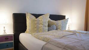 A neatly made bed with floral-patterned pillows and a white comforter. There is a nightstand with a lamp and a multi-drawer cabinet. - Landhaus Valentin (Triberg im Schwarzwald)
