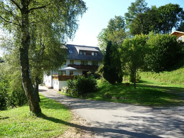 A two-story house with a sloped roof, a balcony with a wooden railing, and a paved driveway leading to the entrance.