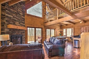 A spacious cabin interior with a stone fireplace, leather sofas, a dining area, and large windows offering views of the surrounding forest. - Family-Friendly Warsaw Cabin w/ Deck & Fireplace! (Warsaw)