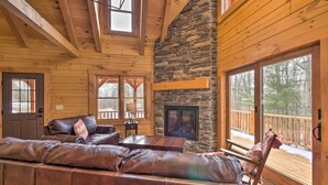 A cozy cabin interior with a stone fireplace, leather sofa, and large windows overlooking a deck.