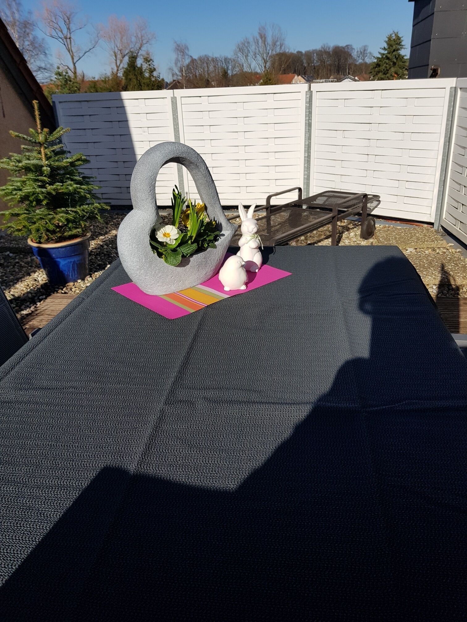 A patio with a gray outdoor table, a stone planter with flowers, and a white rabbit figurine.