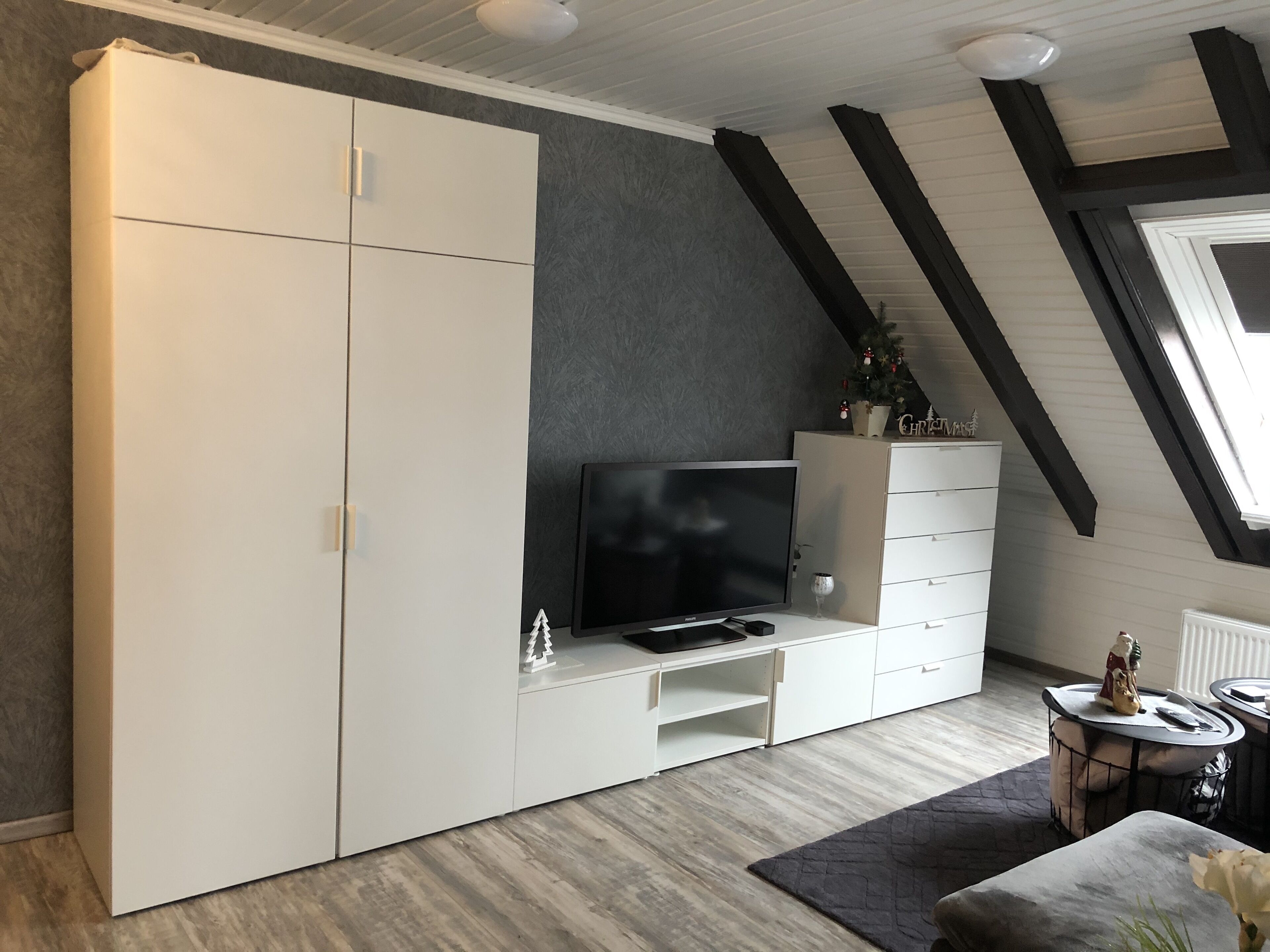 A modern living room with a large white wardrobe, a flat-screen TV on a stand, a small table with a decorative item, and a window with a view of the sky.