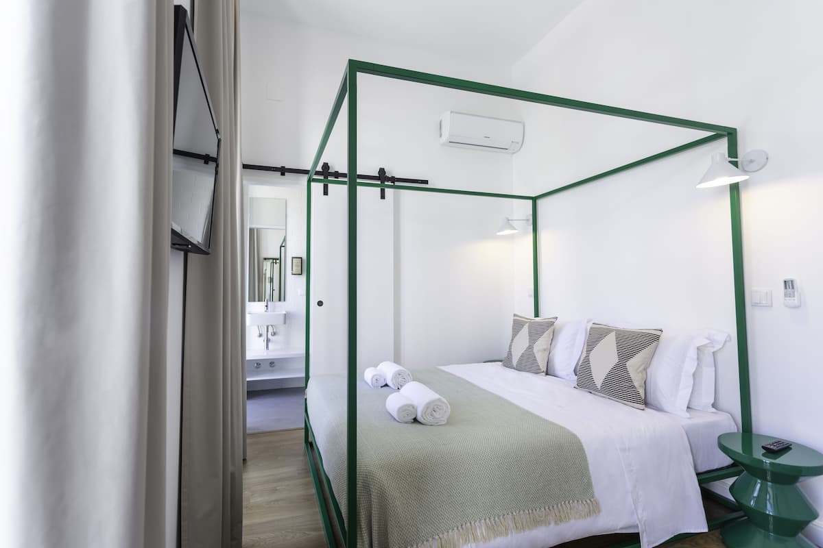 A modern hotel room with a four-poster bed, a green accent wall, a bedside table, and a mirror.