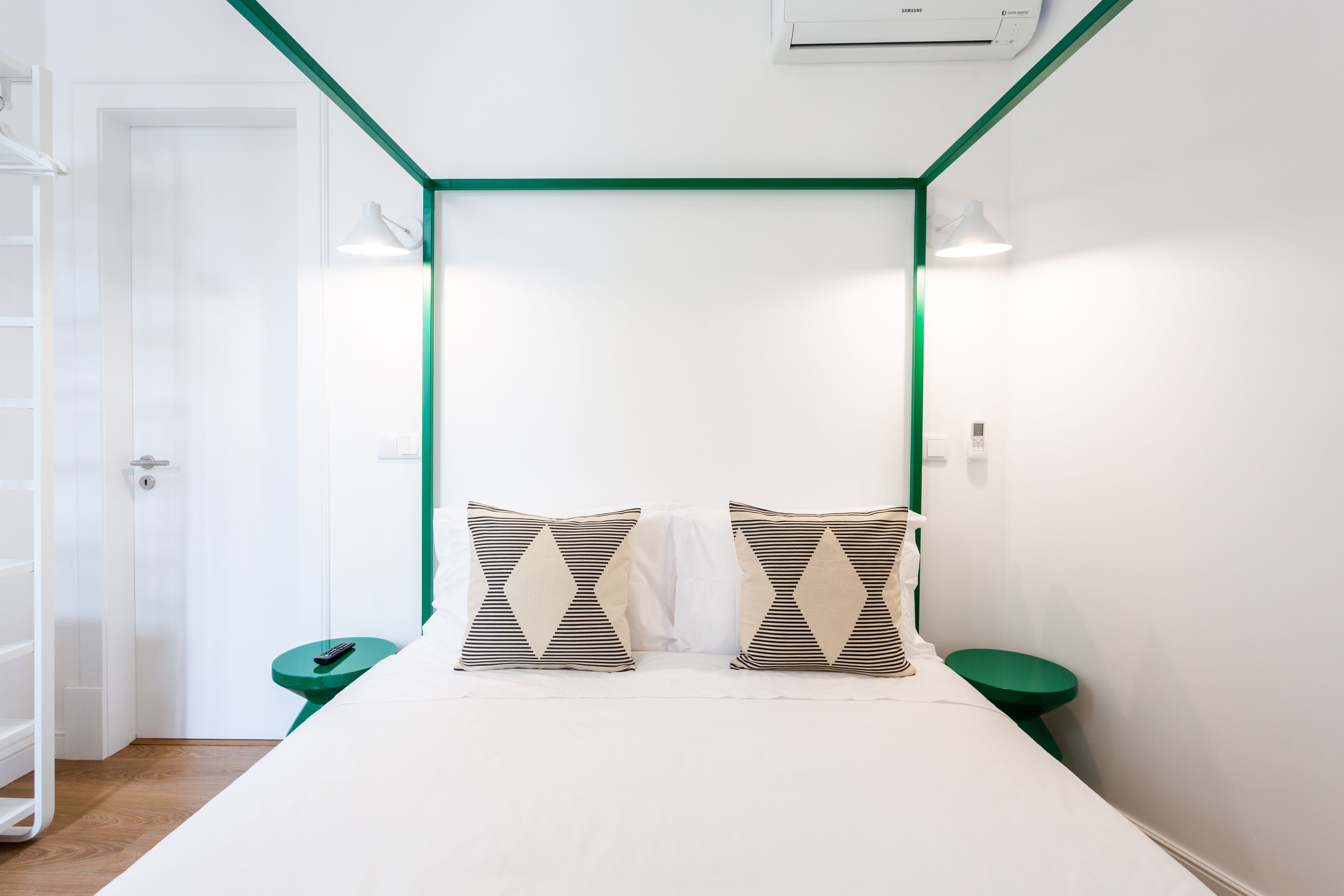 A small, modern room with a bed, two pillows, a green nightstand, a white door, and a green-framed ceiling.
