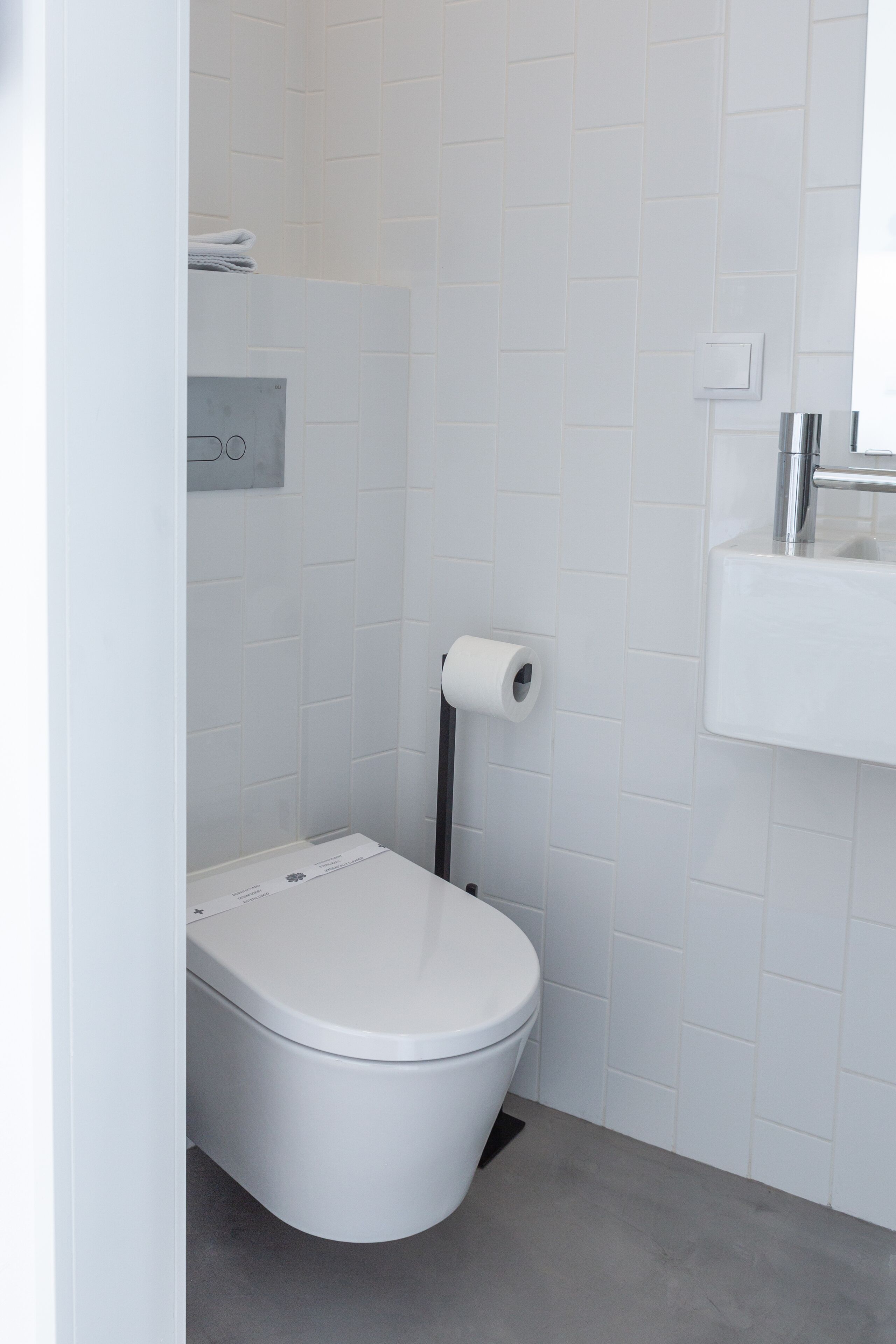 A modern bathroom with white tiles, a white toilet, a paper towel dispenser, and a sink with a mirror.