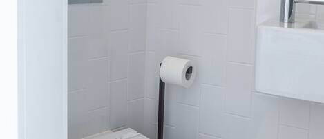 Et moderne badeværelse med hvide fliser, et hvidt toilet, en papirhåndklædeholder og en vask med spejl.