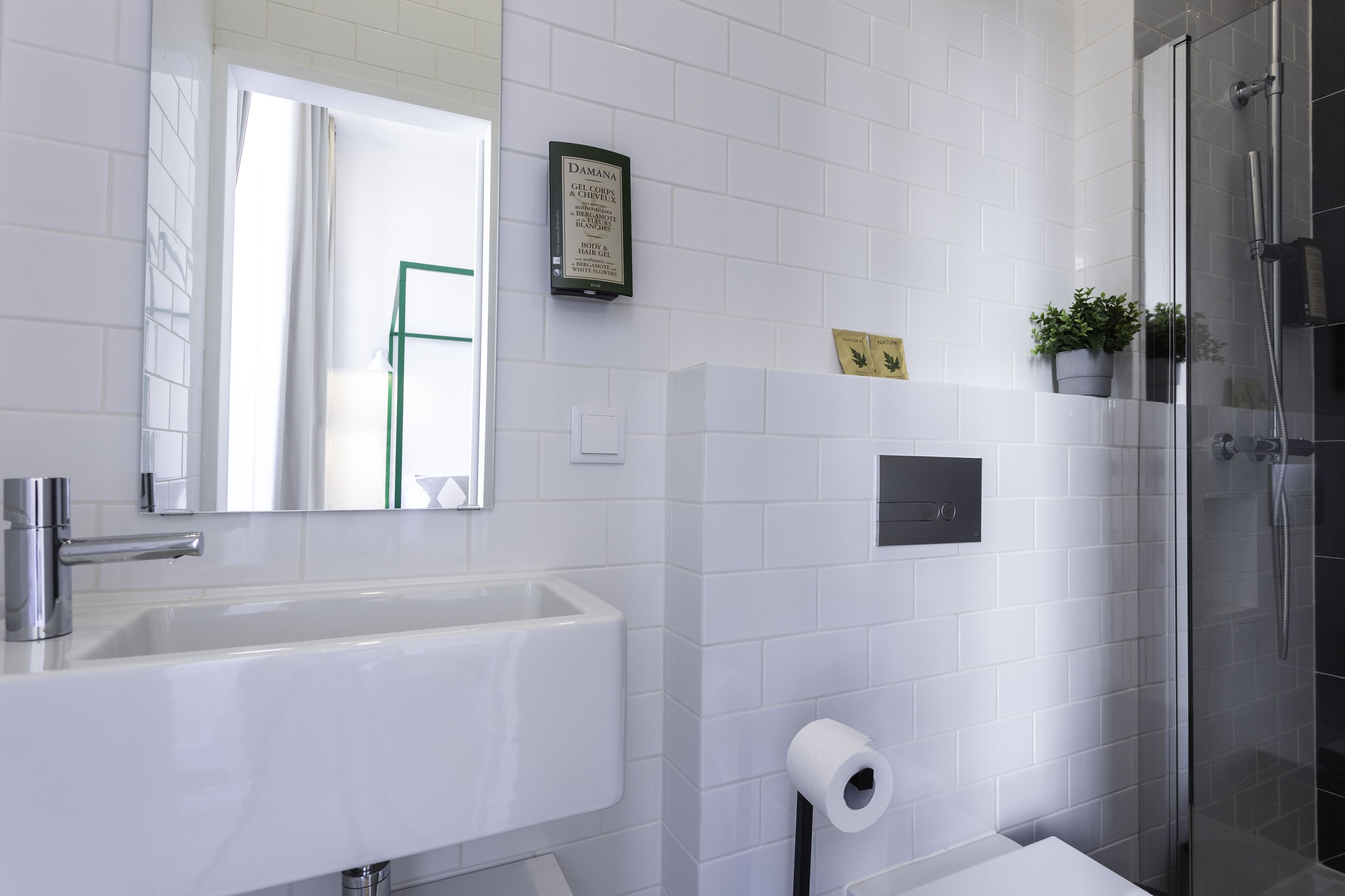 Un baño moderno con un lavamanos, espejo y ducha blancos. Hay un certificado en un marco en la pared y una planta pequeña en el borde de la ducha.