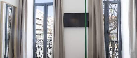 A hotel room with a bed, a TV mounted on the wall, and large windows with curtains.