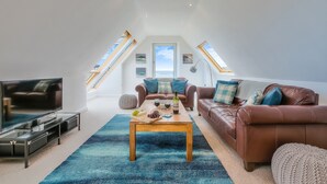 Ein geräumiges Loft mit hohen Decken, einem großen Fernseher, einem Holztisch und einem braunen Ledersofa.
