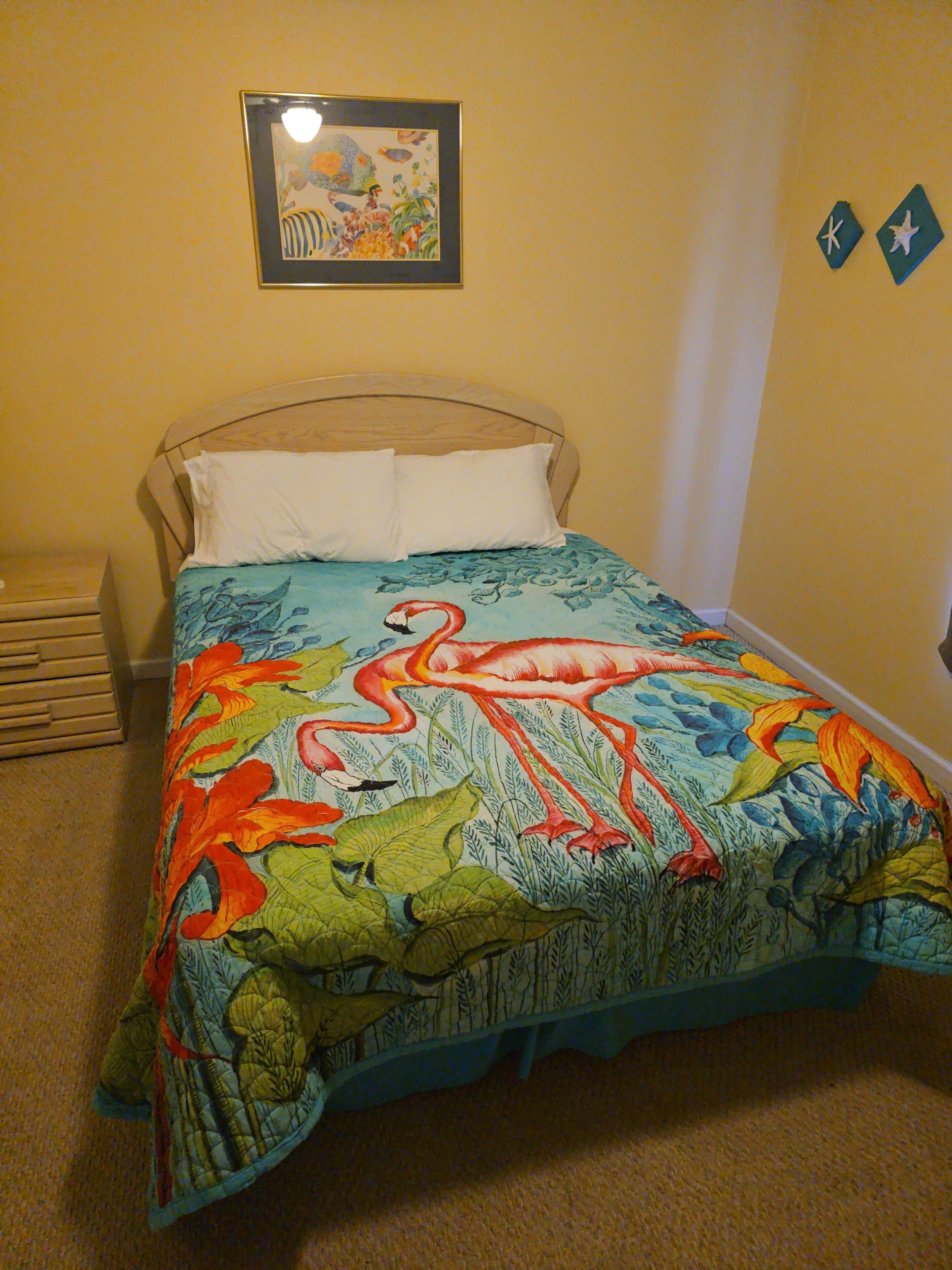 A bed with a colorful floral and animal print bedspread, a wooden headboard, and a small bedside table with a lamp.