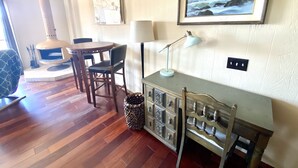 A cozy room with a wooden floor, a round table with chairs, a cabinet with drawers, a lamp, and a framed painting of the sea. - Rarely Available Oceanfront Penthouse (San Diego)