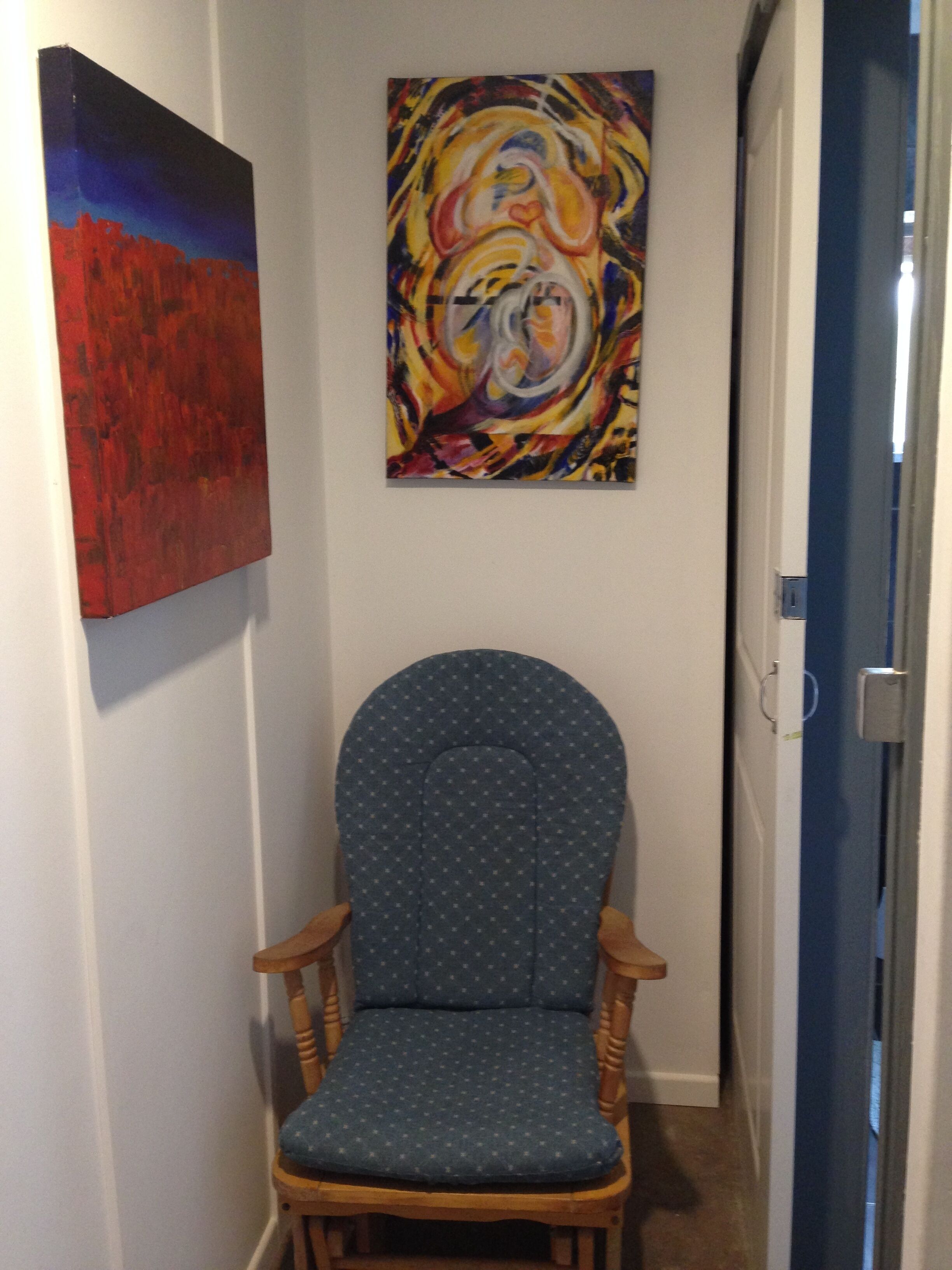 A hallway with a blue upholstered chair, a red abstract painting, and a colorful abstract painting on the wall.
