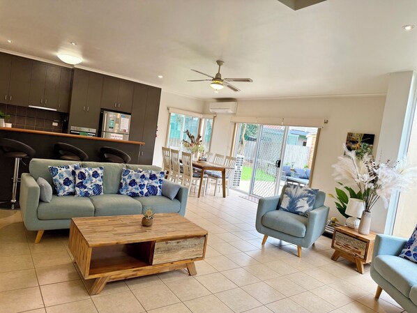 A modern living room with a sofa, armchairs, and a coffee table. There is a kitchen area with cabinets and a dining table. - Whites Retreat (The Esplanade) (Torquay)