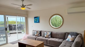 A living room with a sofa, a wooden coffee table, a ceiling fan, a large window, and a view of the outdoors. - Whites Retreat (The Esplanade) (Torquay)
