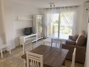 A modern living room with a wooden dining table, white chairs, a brown sofa, and a TV on a white cabinet.