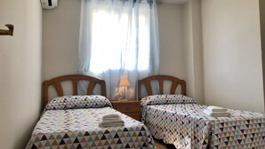 A room with two single beds, a window with sheer curtains, a bedside lamp, and a wall-mounted air conditioner.