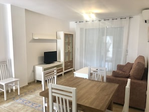 A living room with a wooden dining table, white chairs, a brown sofa, a TV on a white stand, and a glass cabinet. - Ideal apartment for families with playground, pool and garden (Denia)