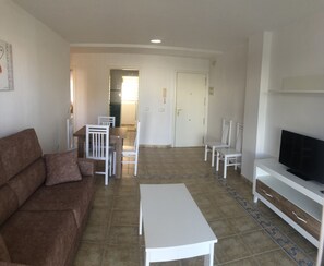 A living room with a brown sofa, a white coffee table, a TV on a wooden stand, and a dining area with white chairs and a table.