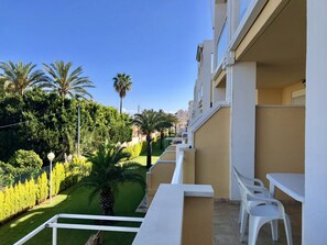 Un balcon avec des chaises blanches et une table, donnant sur un jardin bien entretenu, agrémenté de palmiers et de haies taillées avec soin.