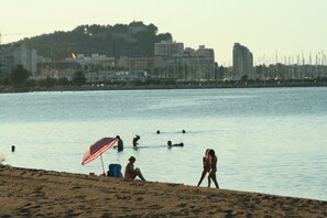 A beach with people swimming and walking, a sandy shore, and a cityscape in the background.