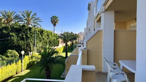 A balcony with white chairs and a table, overlooking a well-maintained garden with palm trees and neatly trimmed hedges.