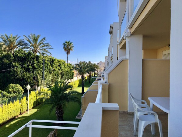A balcony with white chairs and a table, overlooking a well-maintained garden with palm trees and neatly trimmed hedges. - Ideal apartment for families with playground, pool and garden (Denia)