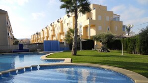 A swimming pool with a curved edge, surrounded by a well-maintained lawn and palm trees. In the background, there is a row of yellow buildings with balconies.