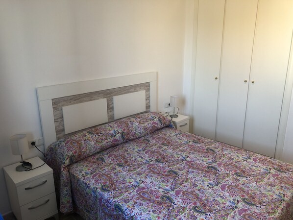 A bedroom with a bed, a nightstand, a lamp, a wardrobe, and a drawer unit.
