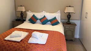 Una cama bien hecha con sábanas blancas y almohadones decorativos, dos mesitas de noche con lámparas, y una colcha estampada.