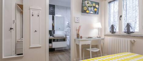A bedroom with a bed, a desk, a chair, a window with curtains, a bathroom with a sink and mirror, and a door leading to another room.