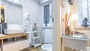 A modern bathroom with a white sink, vanity, and toilet. There is a window with a lace curtain, a shelf with decorative items, and a glass table with a lamp and candles. - B&B Pensieri D'Autore (Campobasso)