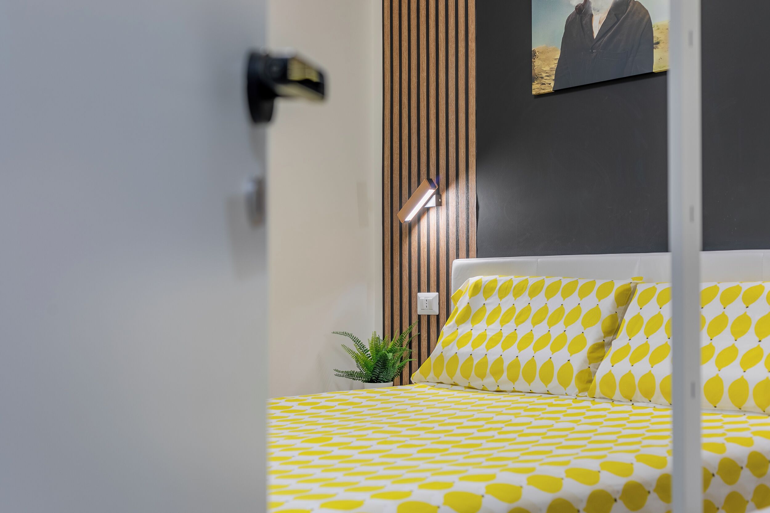 A neatly made bed with a yellow and white patterned bedspread, a small potted plant, and a wall-mounted light fixture.