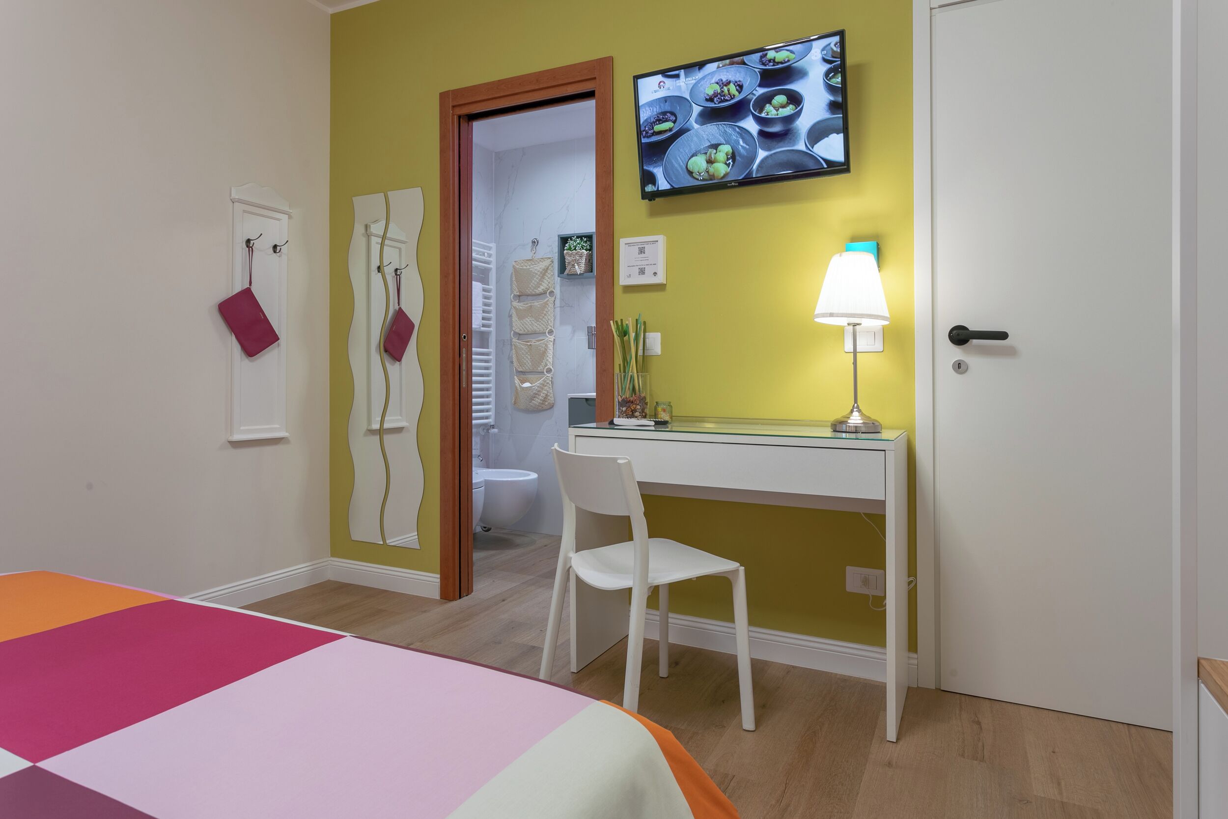 A hotel room with a bed, a desk, a chair, a TV, and a bathroom visible through an open door.