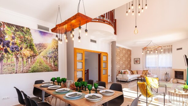 Een ruime eethoek met een lange houten tafel voor zes personen, omringd door zwarte stoelen. Op de muur hangt een groot schilderij van een wijngaard, en de kamer heeft een modern design met een trap naar een hoger niveau.