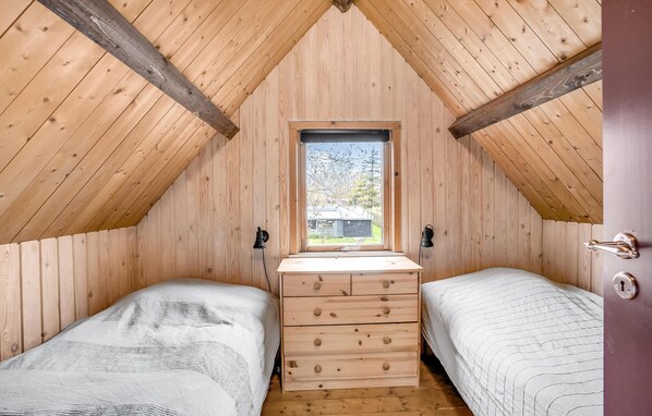 Un pequeño dormitorio en el ático, con paredes de madera, dos camas, un aparador central y una ventana que ofrece vistas al exterior.