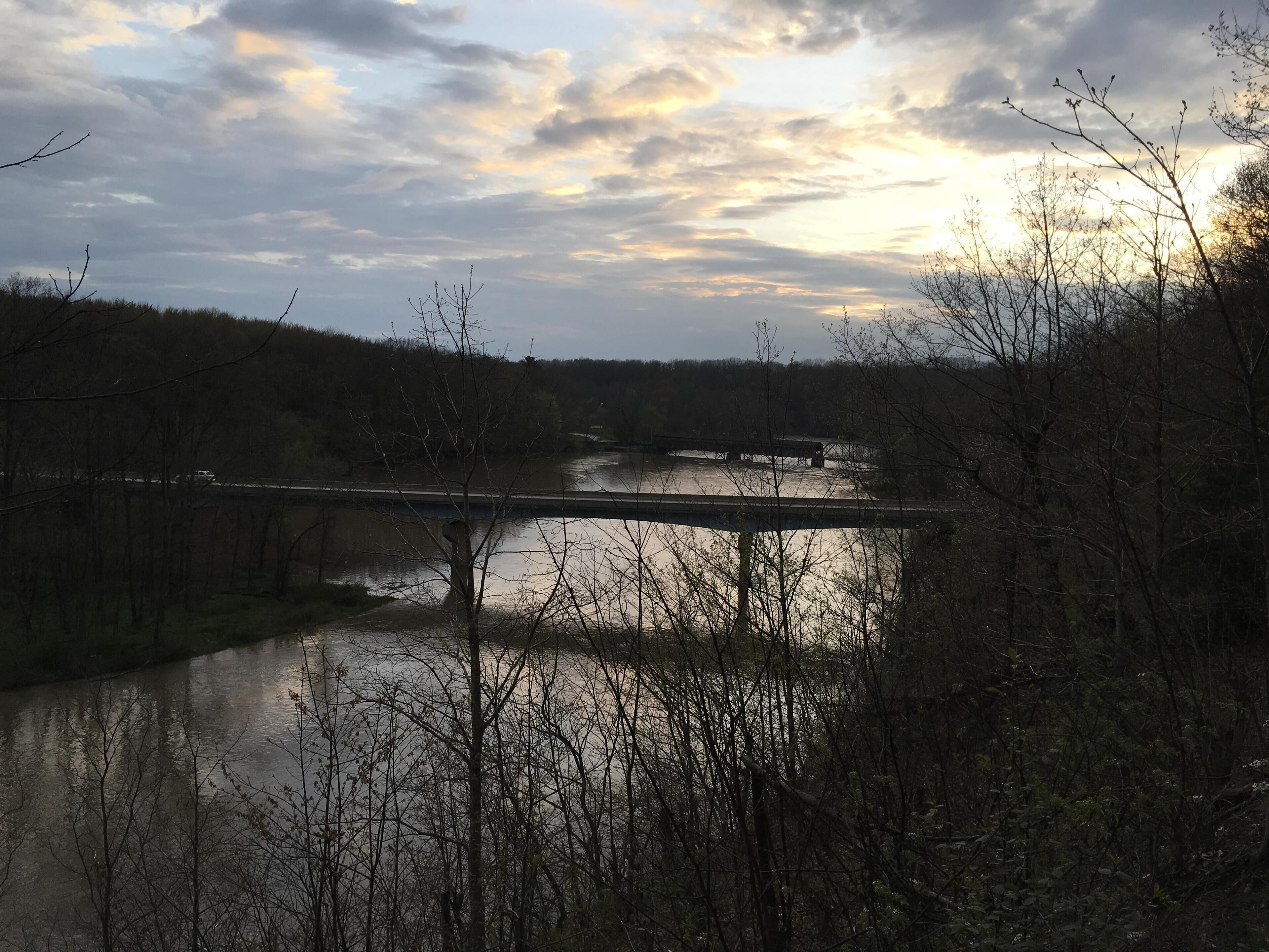Une rivière traversée par un pont, bordée d’arbres et surplombée d’un ciel nuageux.