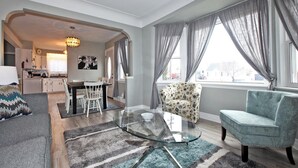 Ein modernes Wohnzimmer mit einem Glastisch, einem gemusterten Sessel und einem hellblauen Sofa. Durch einen offenen Durchgang ist eine Küche erkennbar, ebenso ein Essbereich mit Tisch und Stühlen.