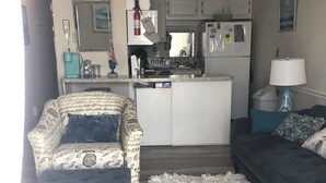 Una sala de estar con sofá, reposapiés y alfombra. Se observa una zona de cocina con armarios y electrodomésticos.